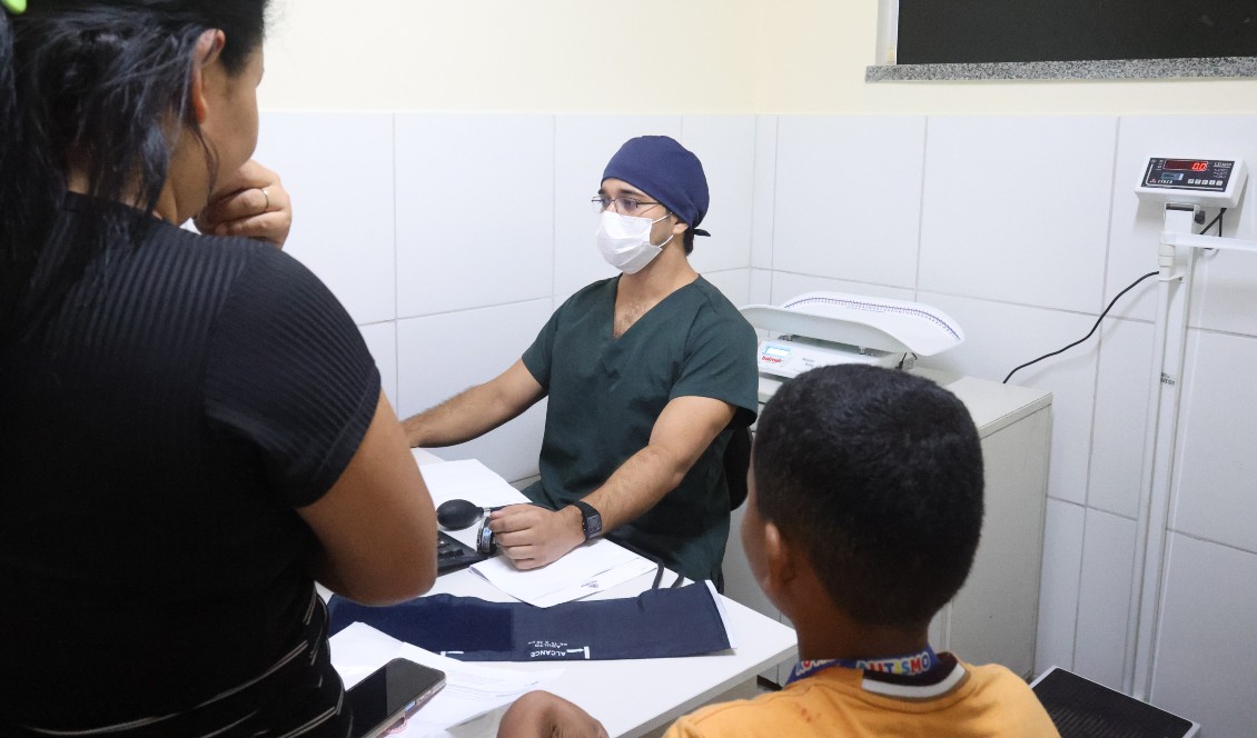 mãe e filho durante uma consulta. Eles estão de costas na frente de um médico que está de máscara